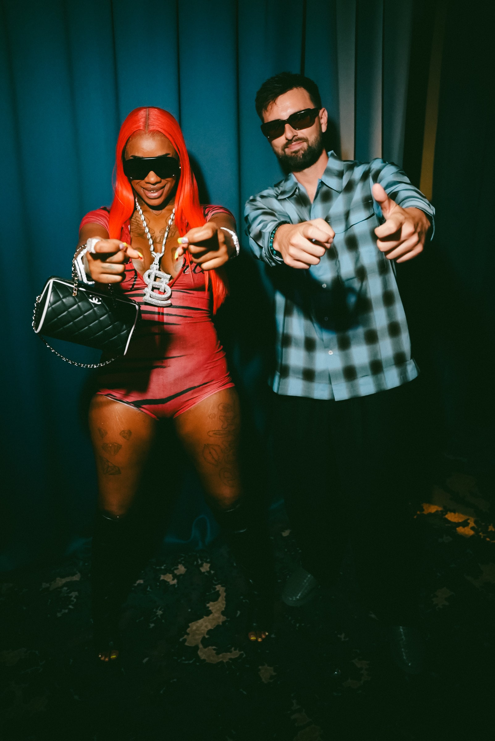 GATANO and Sexyy Red backstage against a blue curtain, both pointing at the camera.